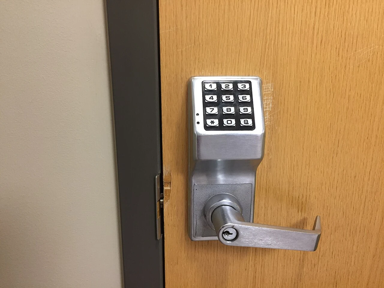 A brushed steel keypad lever lock with a twelve button pad and a key cylinder below the handle, mounted on a wood door