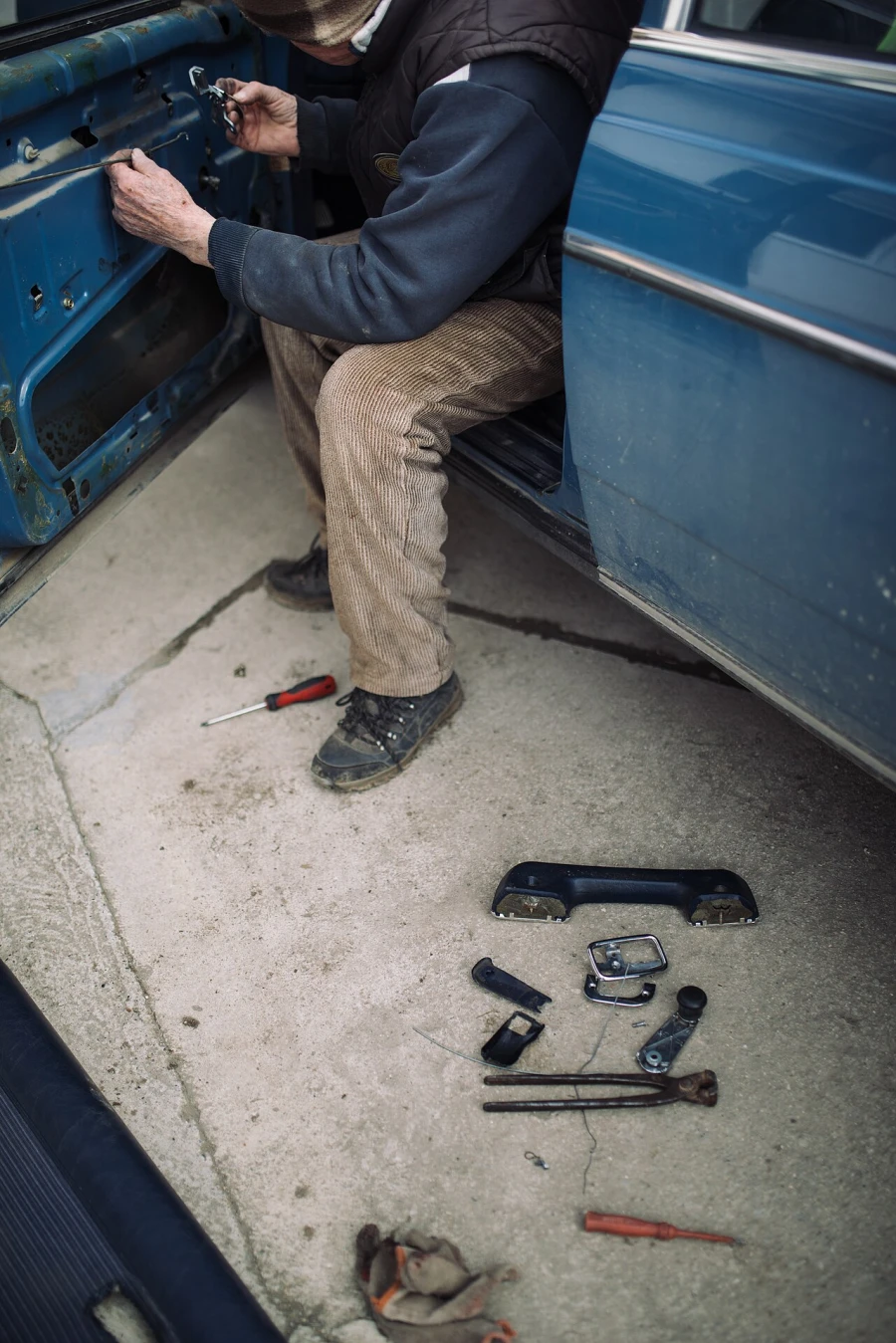 A mechanic sits in an open car door with the panel removed, holding a thin metal lock rod, with handles and tools on the ground