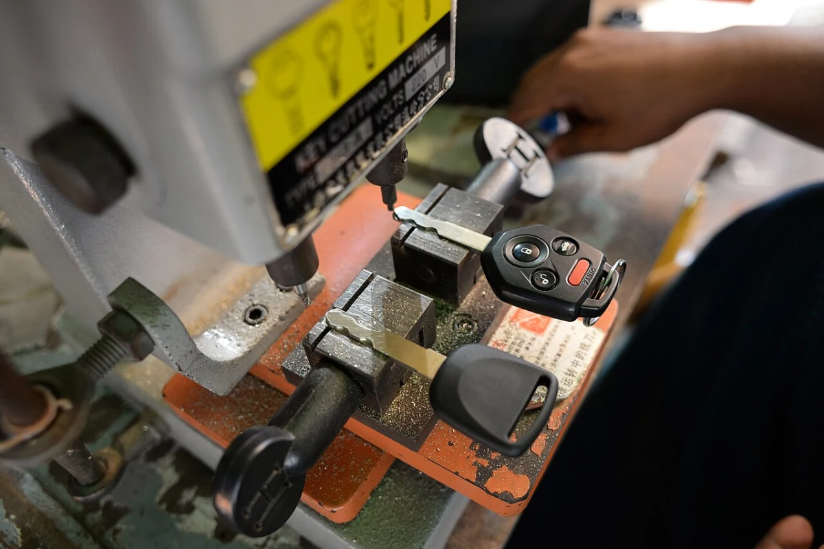 A remote head car key clamped in a key cutting machine next to a second remote key, with a hand on the machine