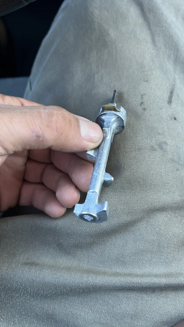 A hand holding an ignition lock cylinder pulled from a steering column, with the key slot at the top and the cam on the bottom