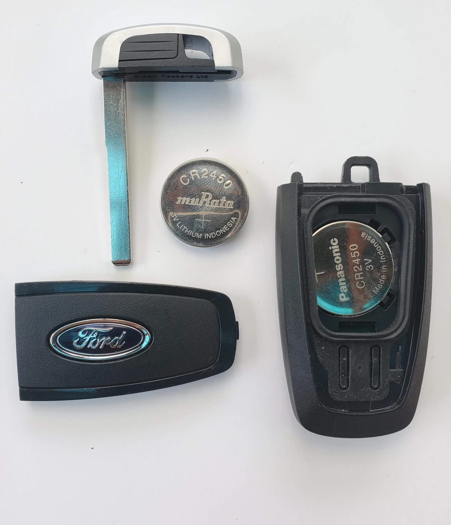 A Ford smart key fob opened up, showing its CR2450 coin cell battery, the removed back cover, a spare battery and the metal emergency key blade